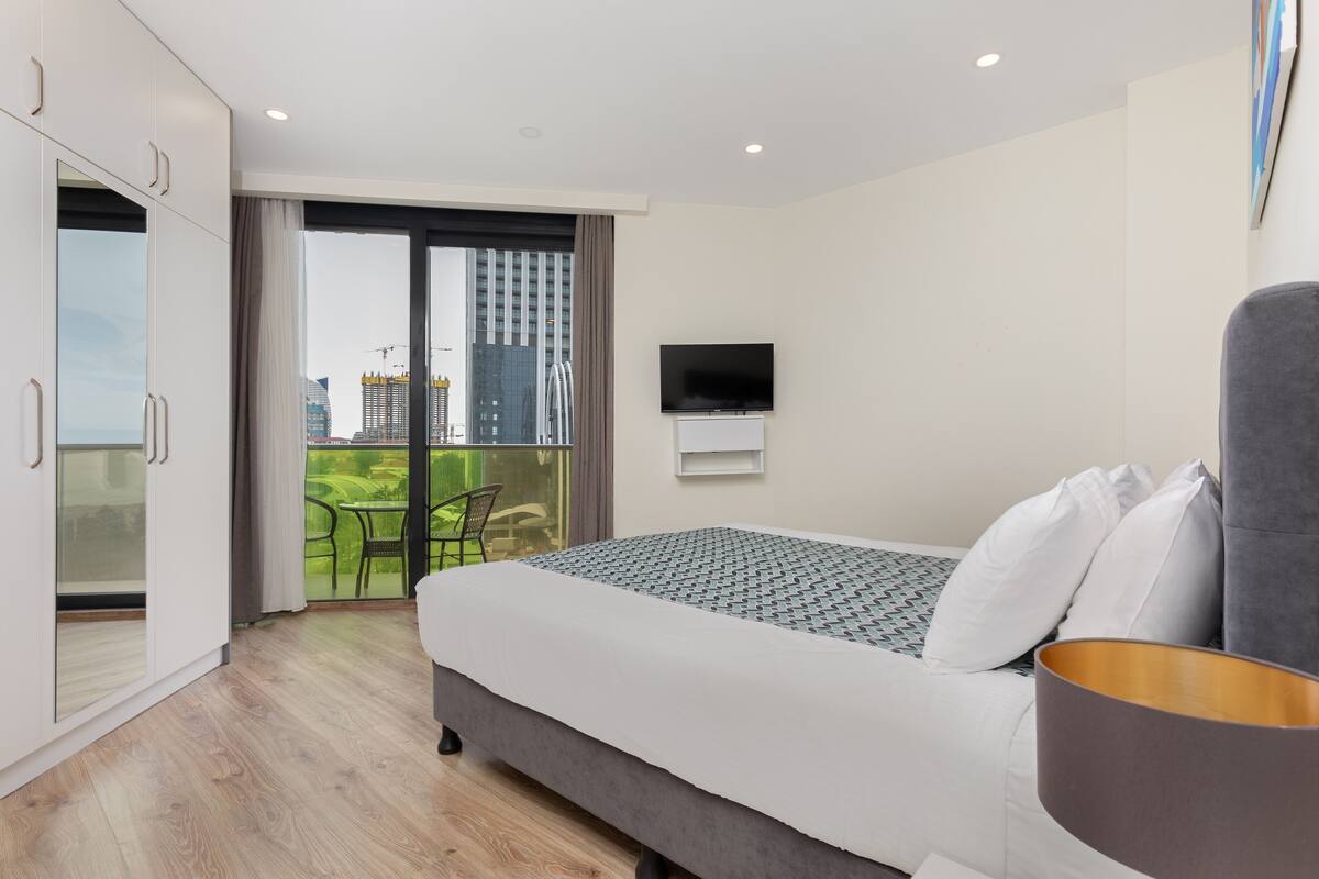 A modern hotel room with a large bed, a flat-screen TV, and a balcony with a city view.