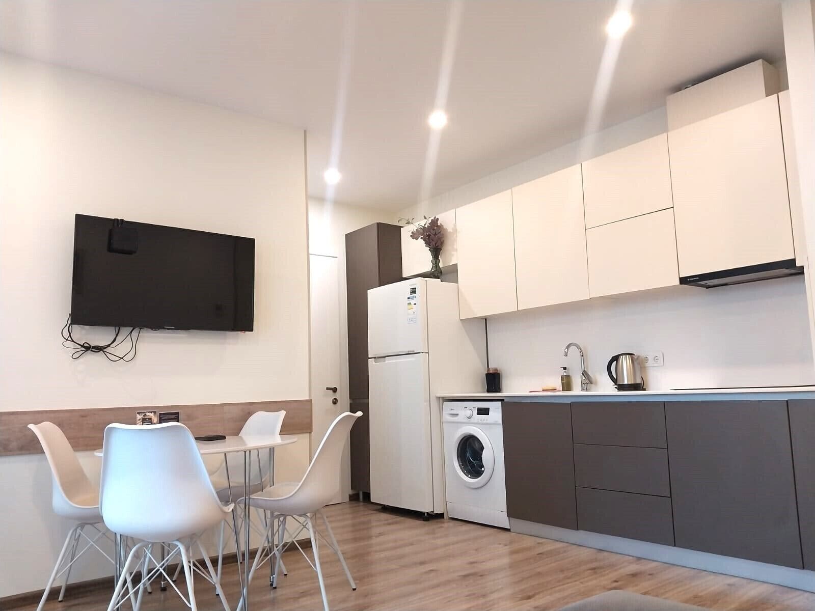 A modern kitchen with a flat-screen TV, a washing machine, and a dining area with chairs.