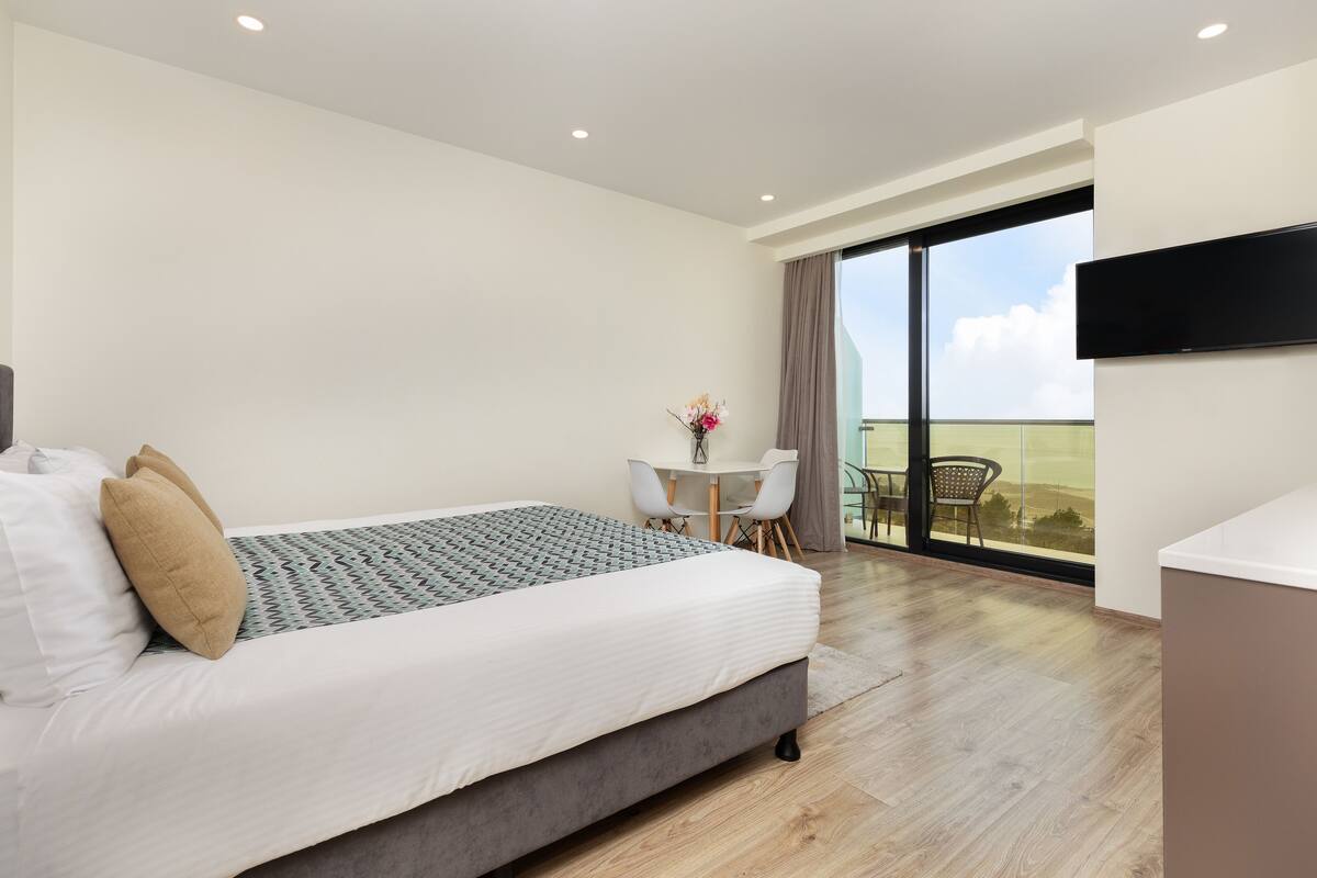 A modern bedroom with a bed, a small table with chairs, a TV, and a balcony with a view.
