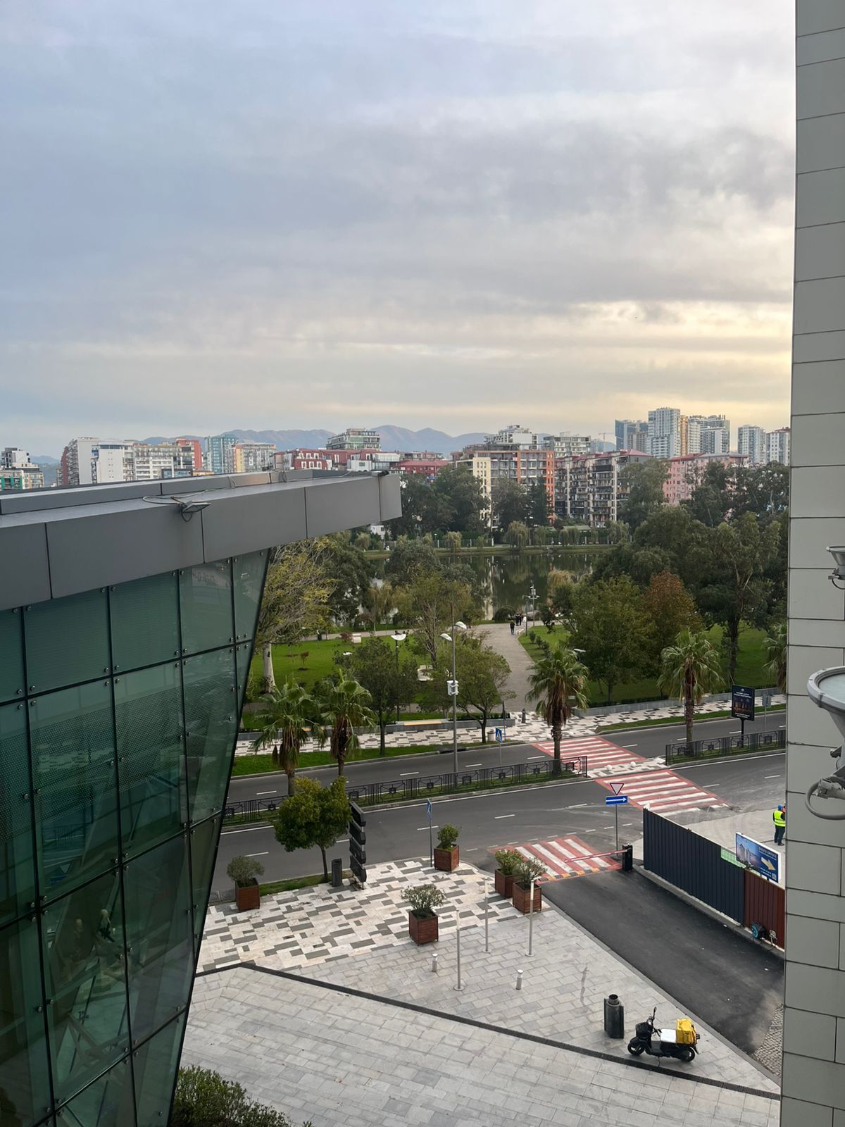 A modern building with a glass facade, a paved plaza with planters, and a view of a cityscape with buildings and a road.