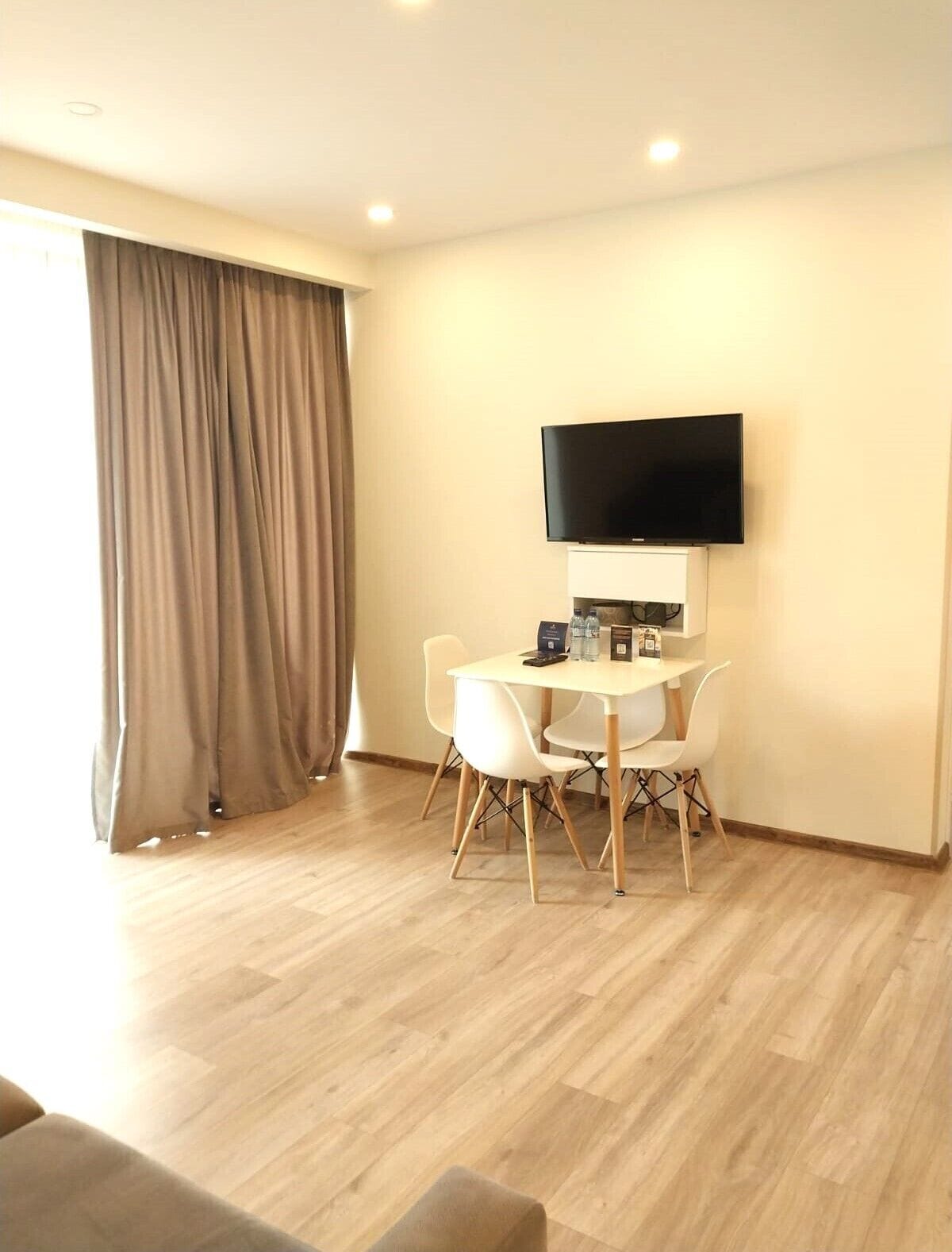 A modern hotel room with a wooden floor, a flat-screen TV mounted on the wall, a small table with chairs, and a large window with curtains.