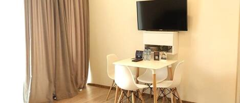 A modern hotel room with a wooden floor, a flat-screen TV mounted on the wall, a small table with chairs, and a large window with curtains.