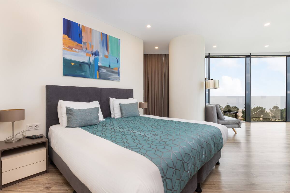 A modern hotel room with a large bed, a nightstand, a chair, and a large window offering a view of the city.