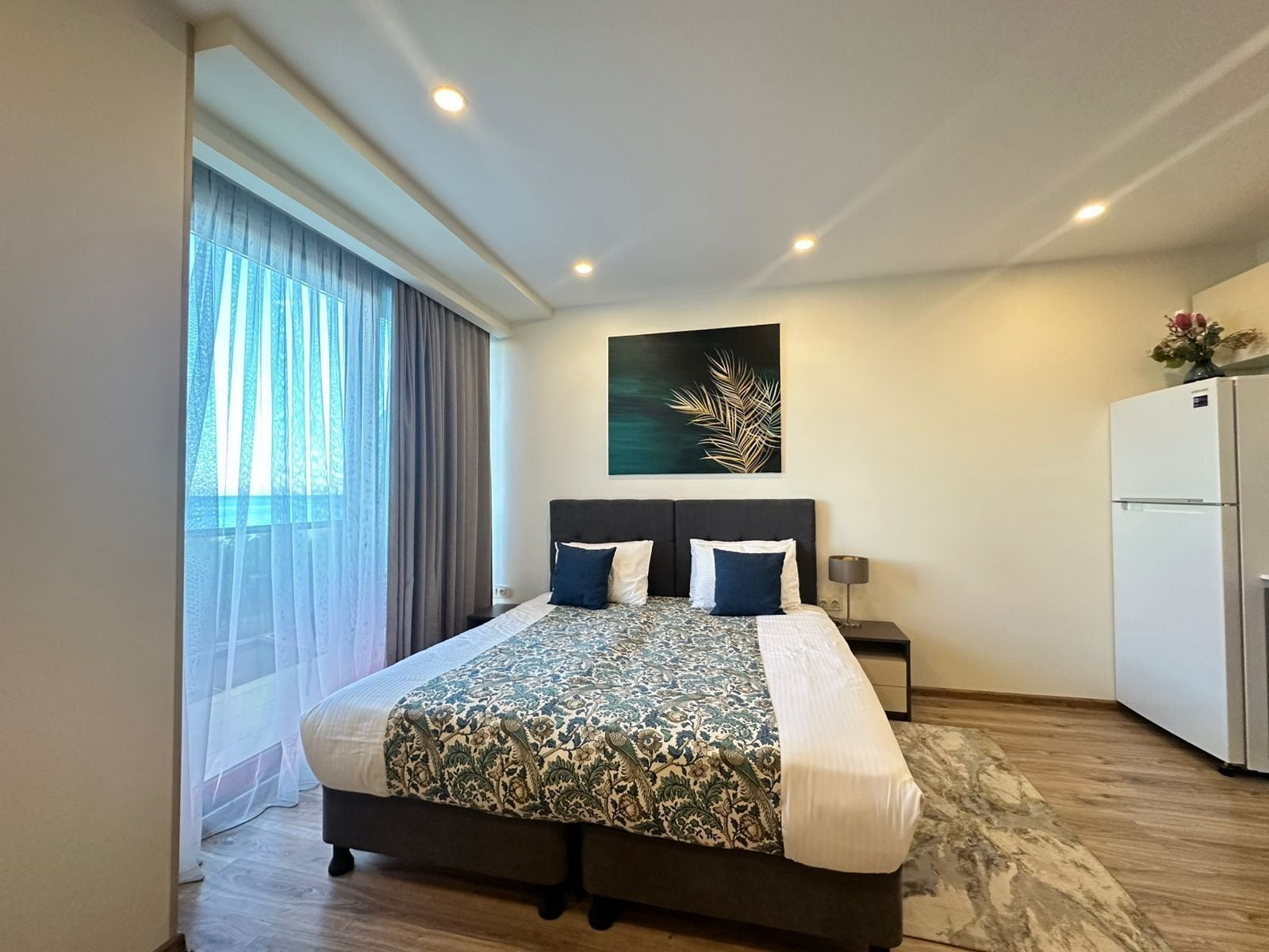 Quarto de hotel moderno com uma cama grande, mesa de cabeceira, obra de arte na parede e frigorífico.