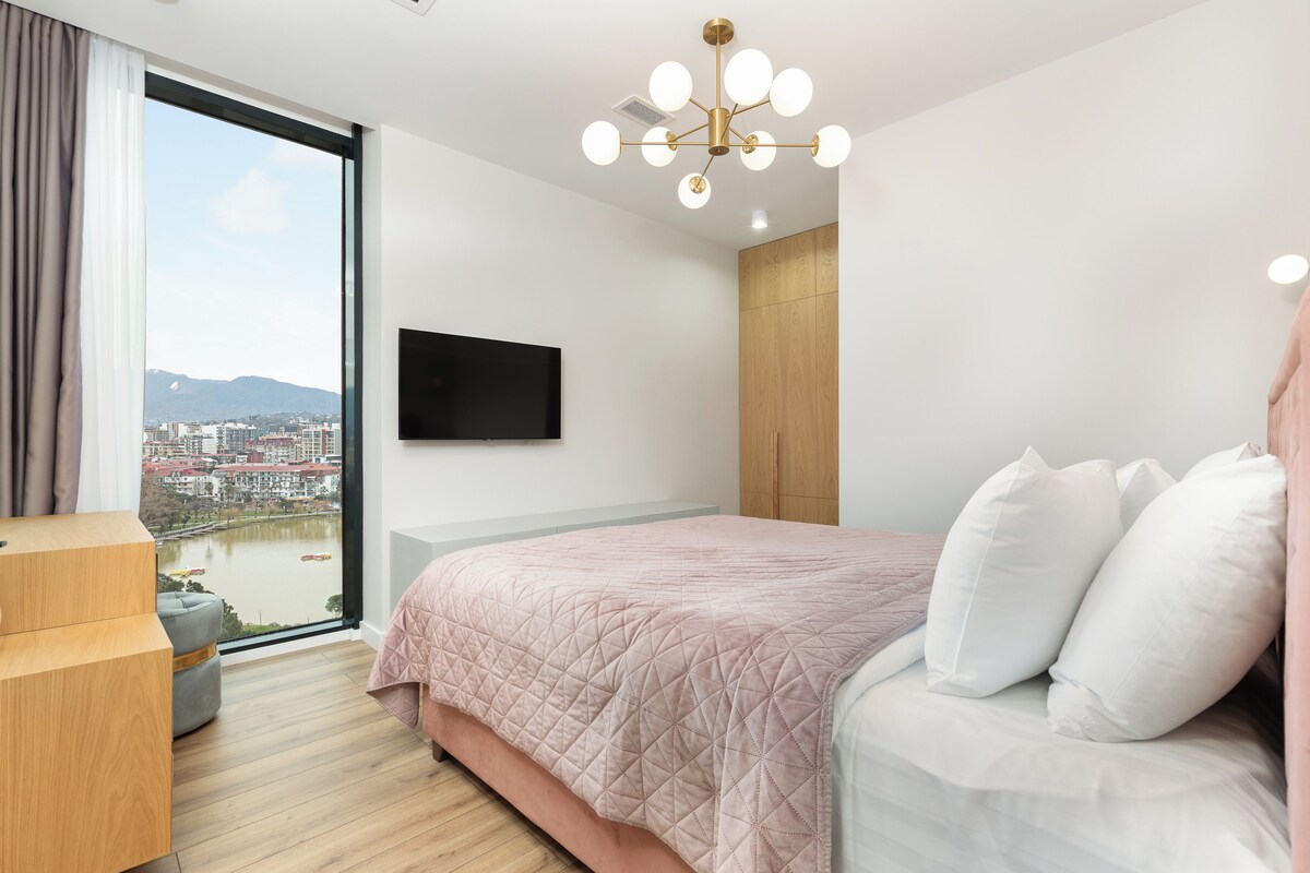 A modern hotel room with a large bed, a TV mounted on the wall, and a view of a cityscape through a large window.