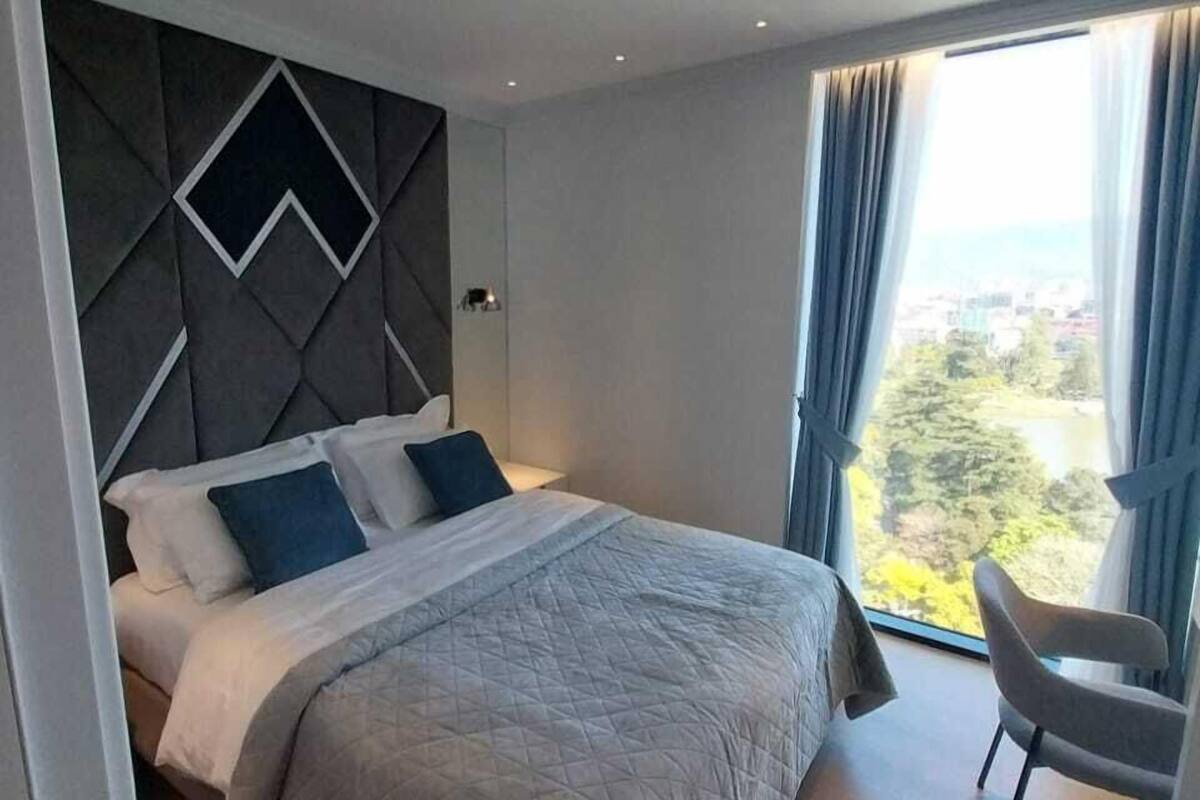 A modern bedroom with a large bed, a chair, and a view of a cityscape through a large window.