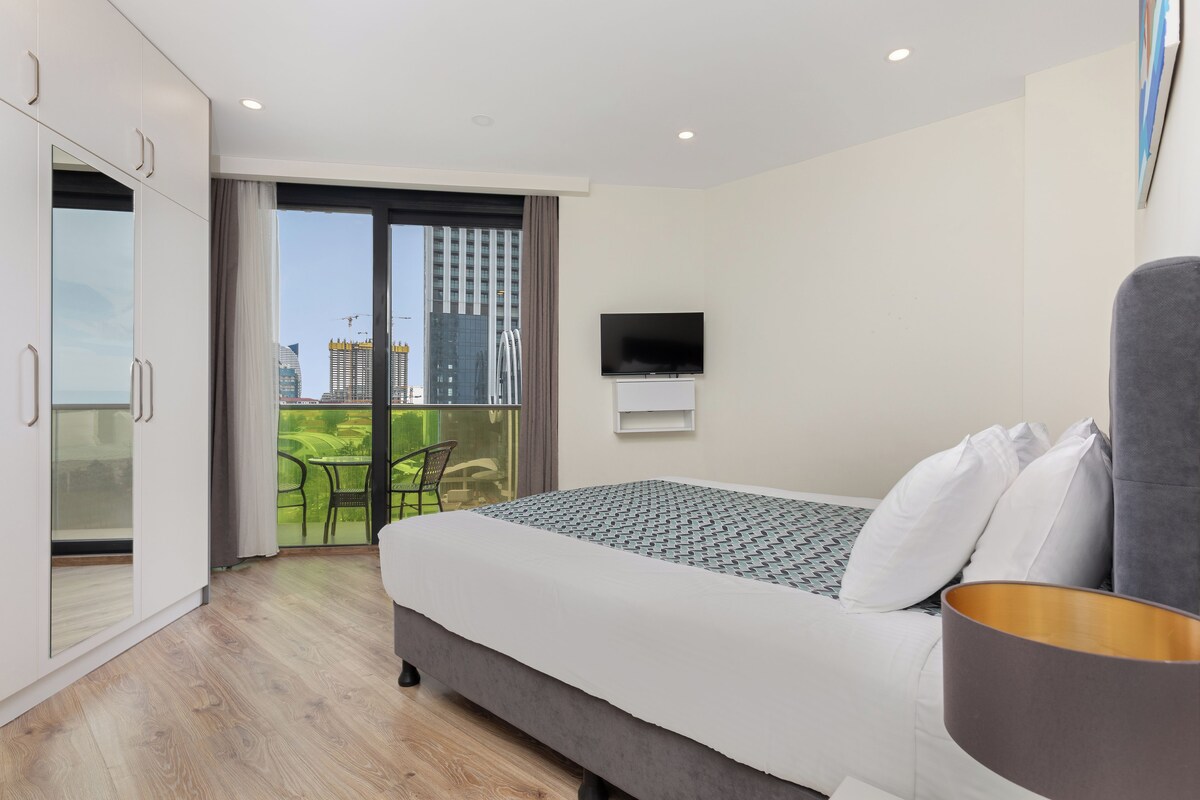 A modern hotel room with a large bed, a flat-screen TV, and a balcony with a view of the cityscape.