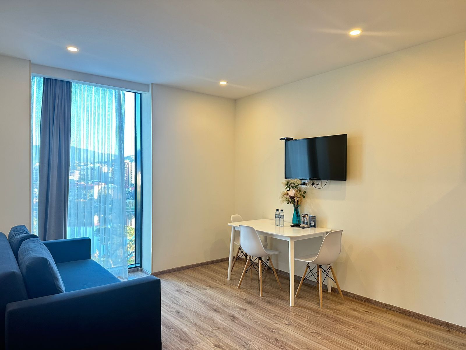 Una sala de estar moderna con un sofá, una mesa de comedor con sillas, un televisor de pantalla plana en la pared y un ventanal con cortinas.
