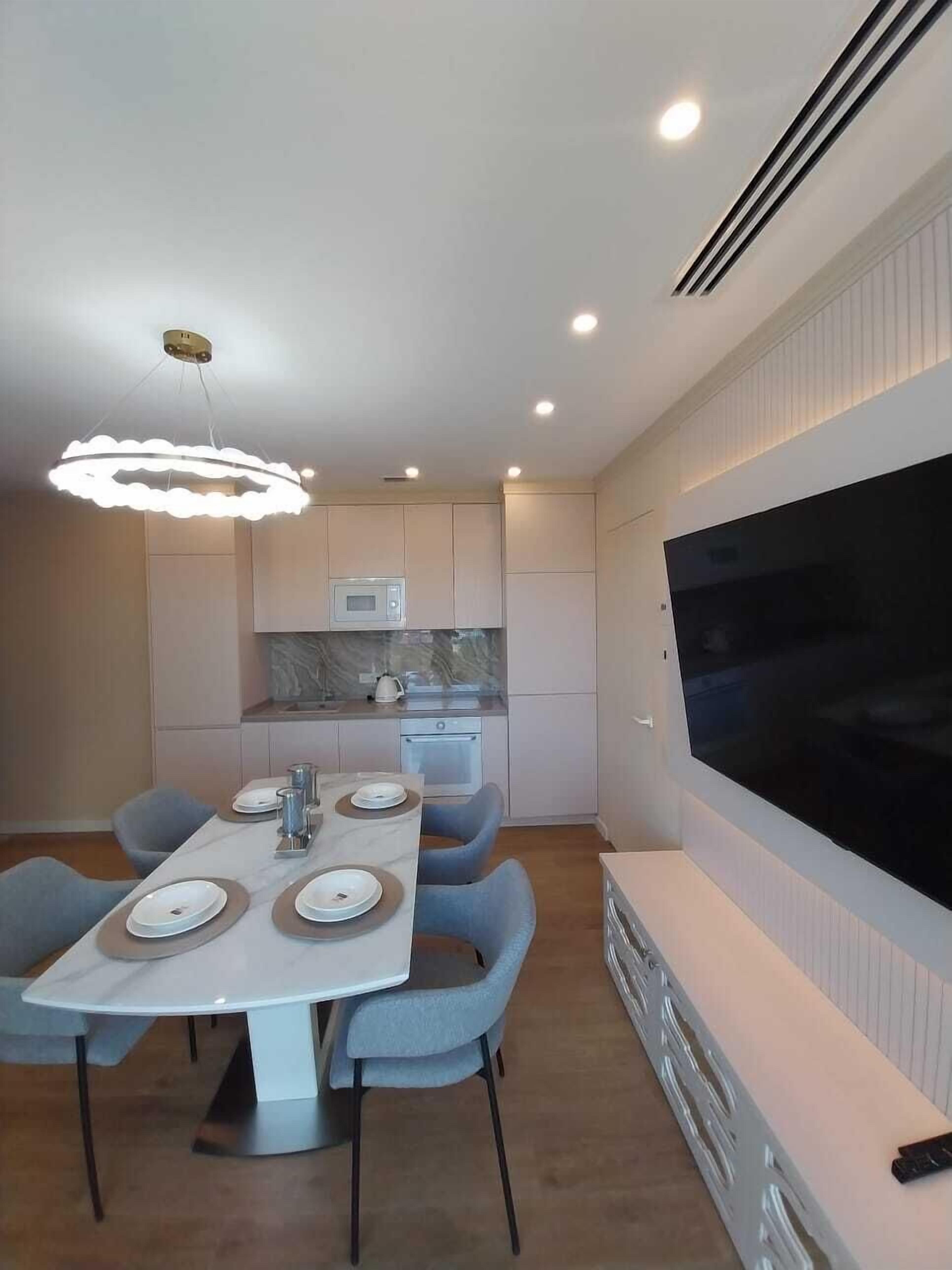 A modern kitchen with a built-in microwave, a dining area with a white table and blue chairs, and a large flat-screen TV mounted on the wall.