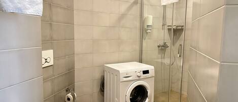 A compact bathroom with a toilet, a washing machine, and a glass shower enclosure.
