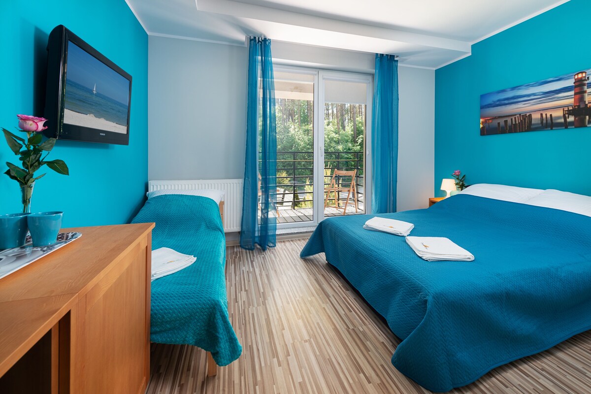 Ein Zimmer mit zwei Betten, einem Holztisch, einer Topfpflanze und Blick ins Freie durch eine Glastür.