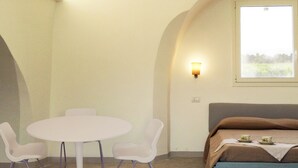 A small, cozy room with a round table and two chairs, a bed with a brown blanket, a wall-mounted light, and a window with a view of greenery. - Casale alle Vigne (Nardò)