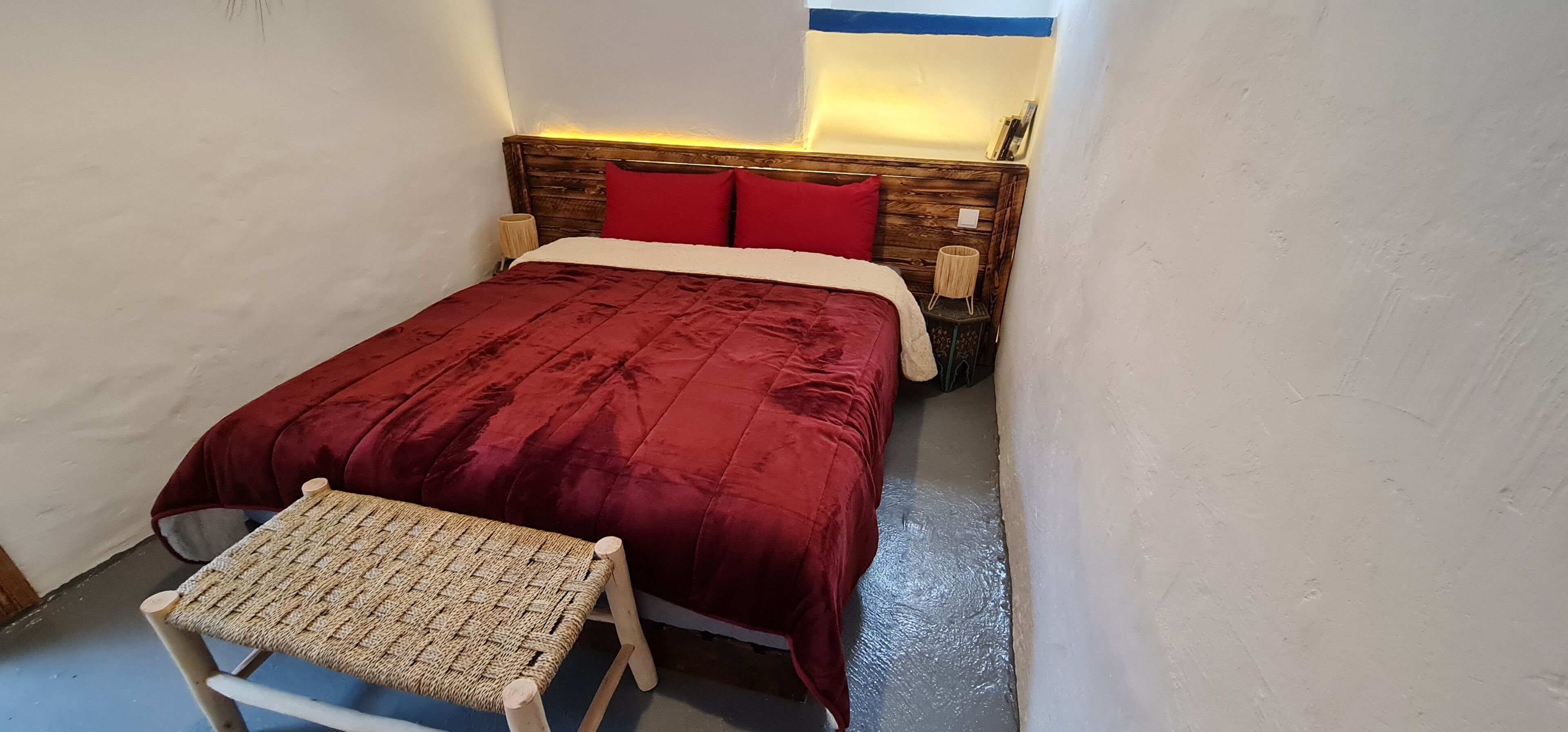Een kleine, gezellige kamer met een houten hoofdbord, een rode sprei en een klein bankje.