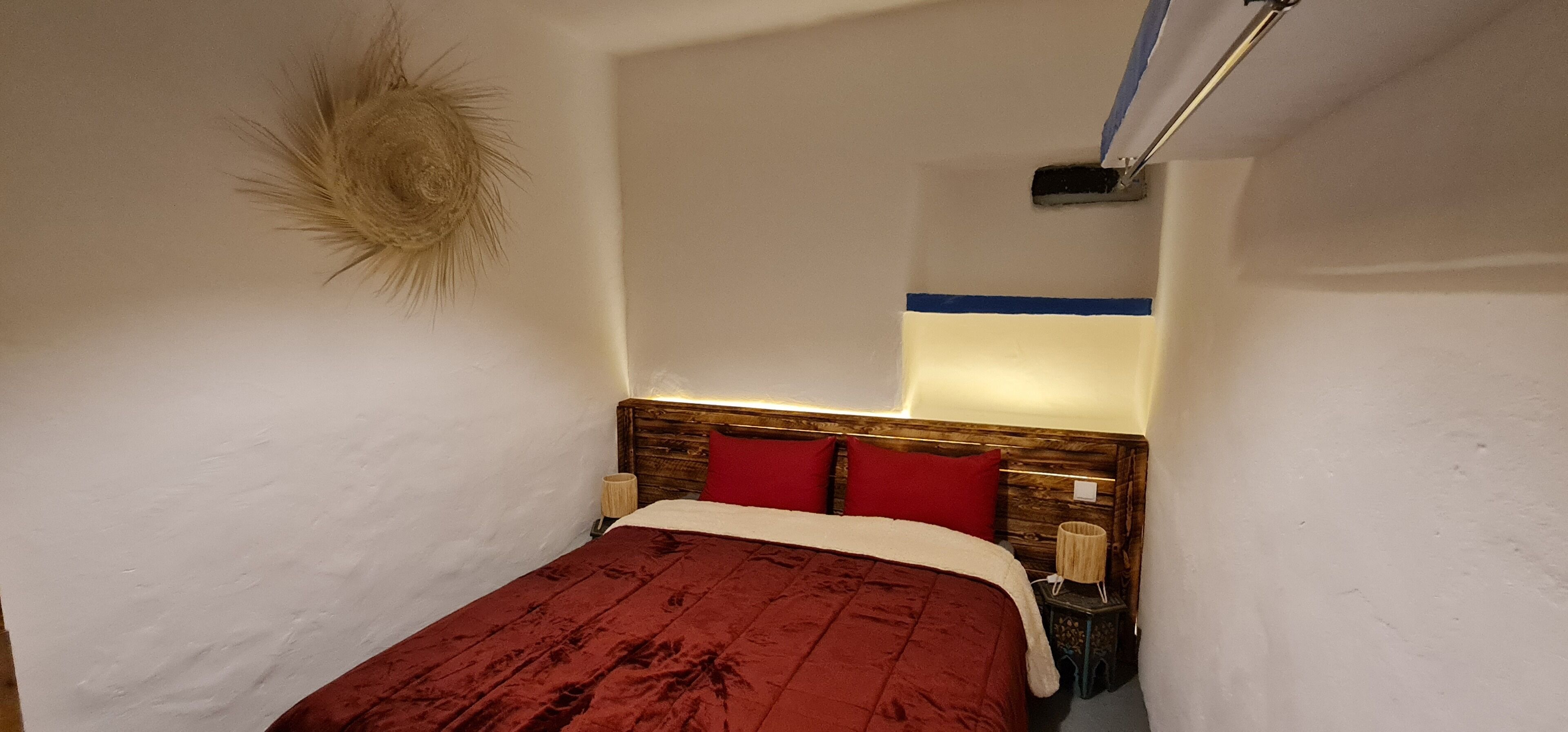 Une petite pièce étroite, avec un lit, une tête de lit en bois et une lampe fixée au mur.