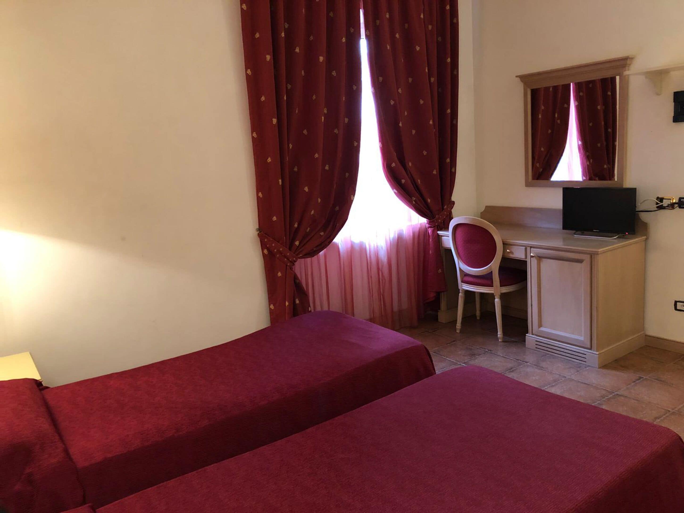 A room with two single beds, a desk with a chair, a television, and a window with curtains.
