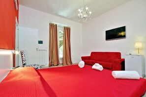 Quarto de hotel com uma cama grande, cortinas vermelhas, um sofá vermelho, uma TV e uma pequena mesa de cabeceira branca.