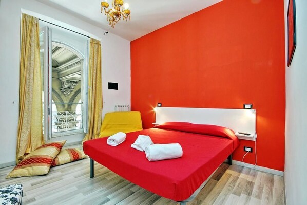 Quarto de hotel com cama vermelha, cadeira amarela e uma janela grande com cortinas.