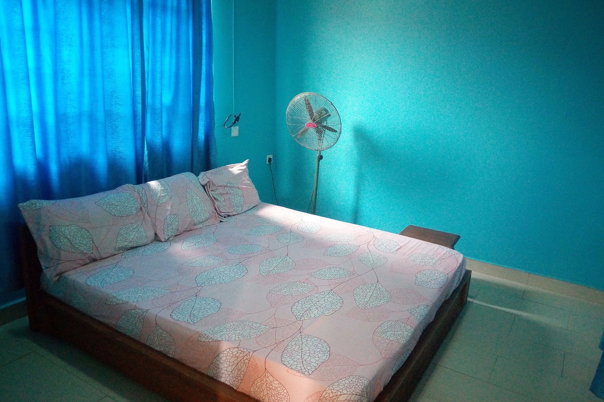 A bedroom with a bed, a standing fan, and a window with blue curtains.