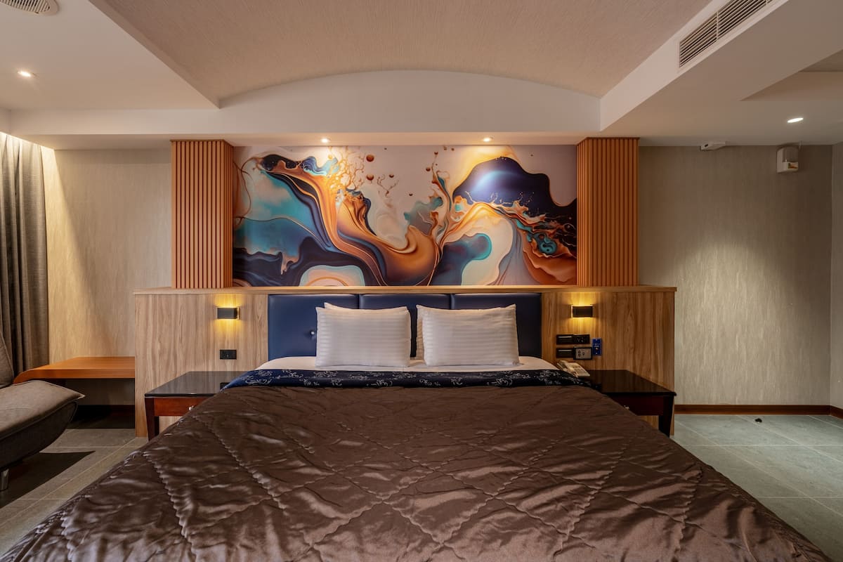 A modern hotel room with a large bed, a headboard with ambient lighting, a wall art piece, and a wooden nightstand.