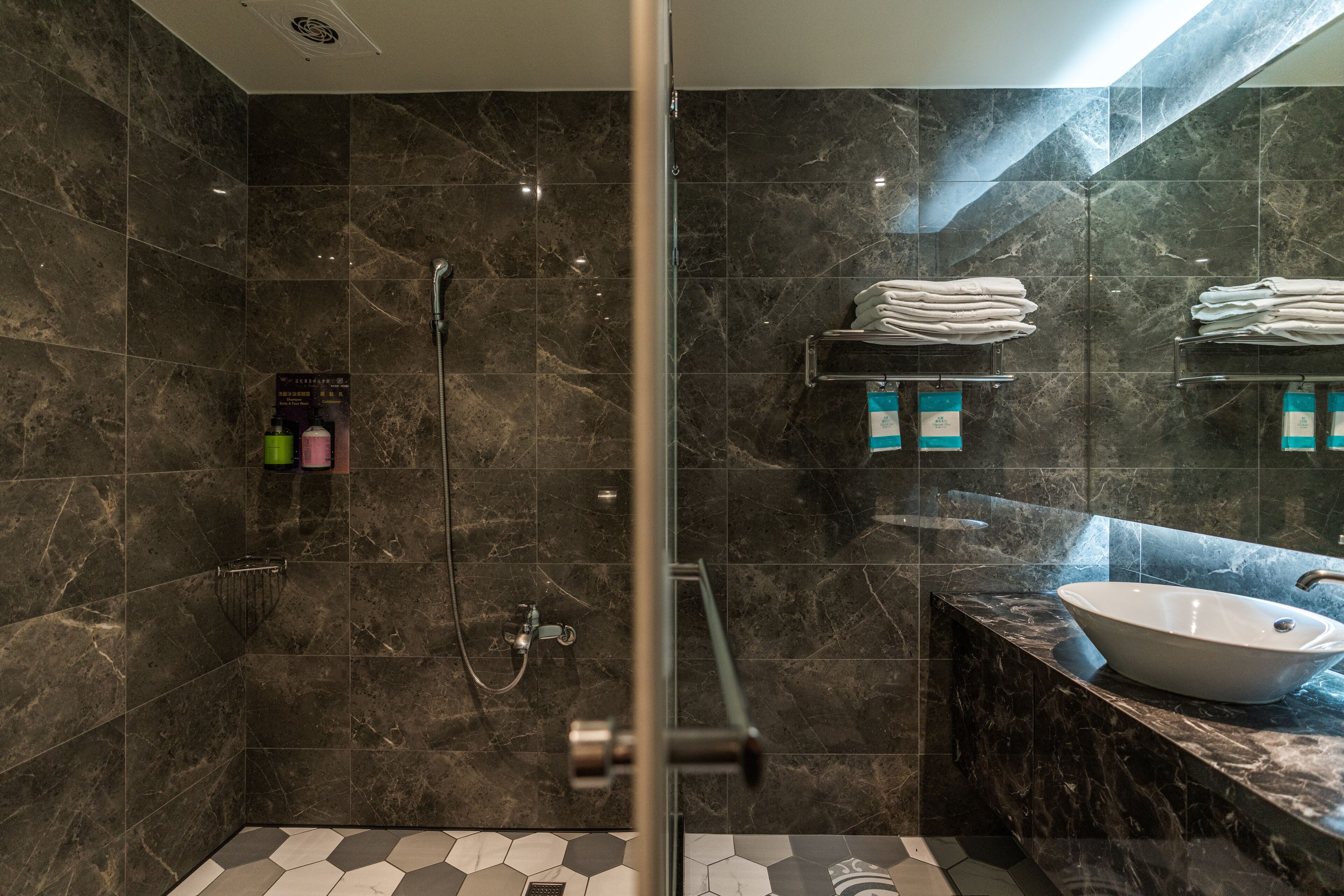 A modern bathroom with dark marble tiles, a glass-enclosed shower, a white sink, and neatly folded towels on shelves.
