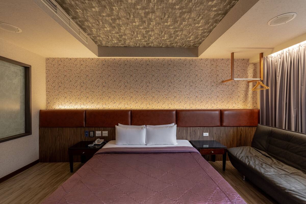 A modern hotel room with a large bed, a sofa, a bedside table, and a headboard with built-in lighting.