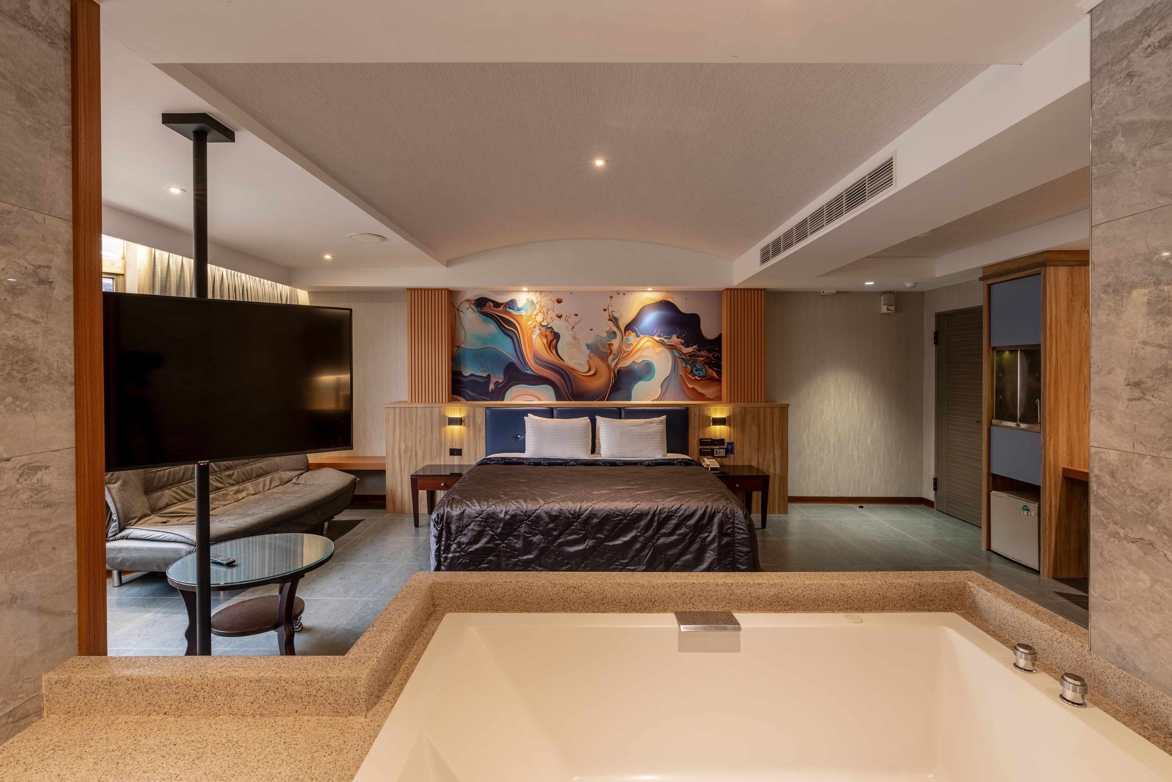 A modern hotel room with a large bed, a sitting area with a sofa and a glass coffee table, a large artwork on the wall, and a bathtub.