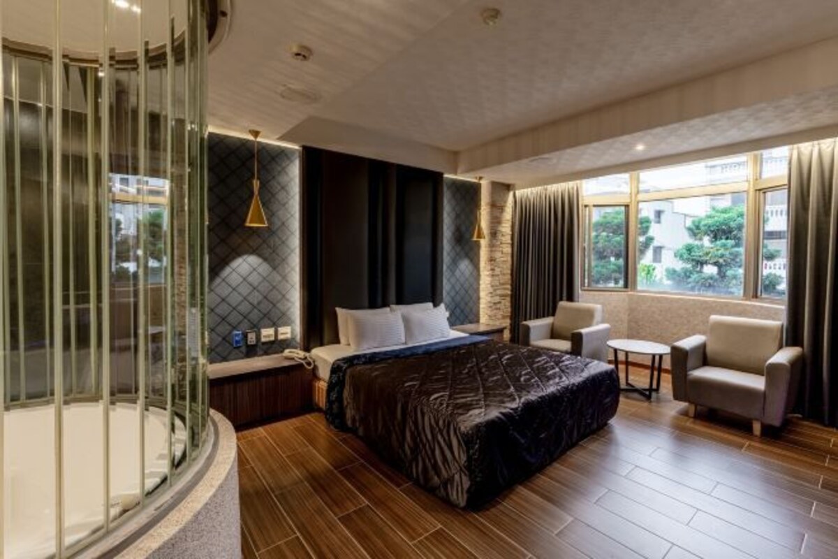 A modern hotel room with a large bed, a seating area with two chairs, a small table, and a glass-enclosed balcony.