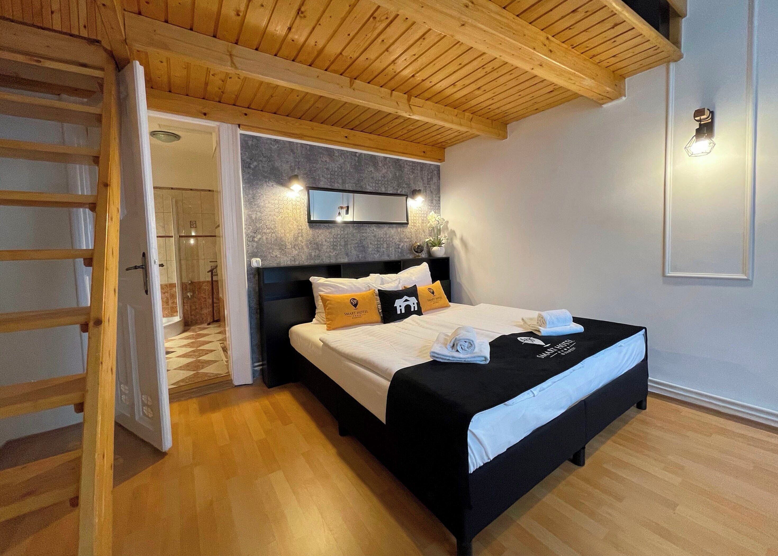Ein modernes Hotelzimmer mit Holzboden, einem großen Bett mit schwarz-weißer Bettwäsche, einem kleinen Nachttisch und einer Wandleuchte.