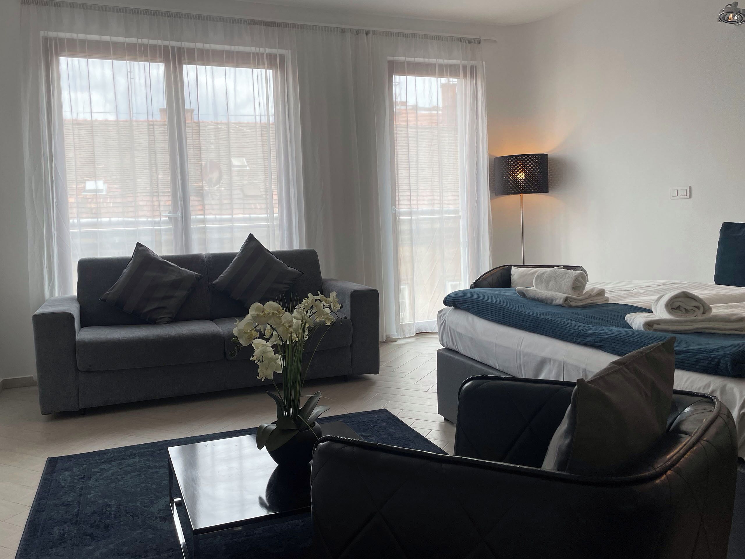 Una habitación de hotel moderna con un sofá gris, una cama con ropa de cama azul, una lámpara de pie y un jarrón con flores blancas.