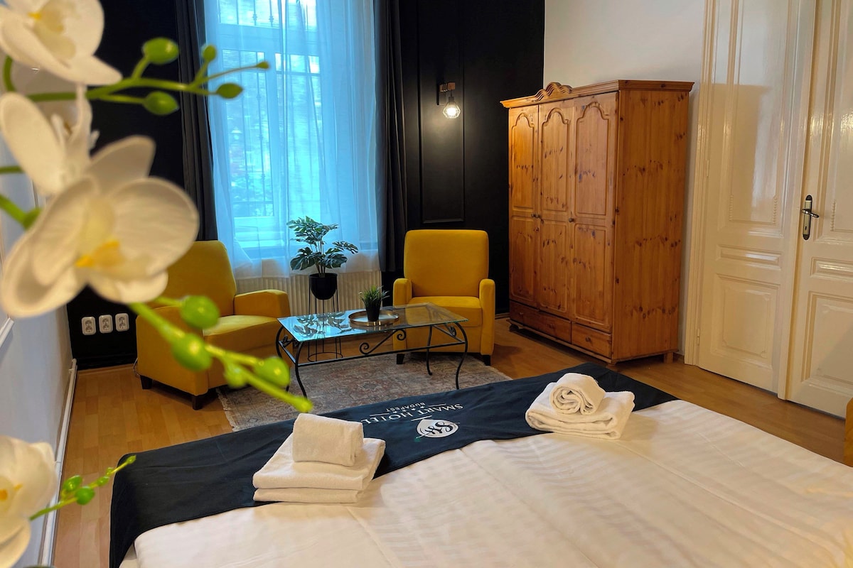 A hotel room with a bed, a yellow armchair, a wooden wardrobe, and a small table with a plant.