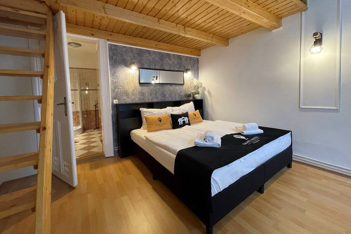 A modern hotel room with a wooden ceiling, a large bed with black and white bedding, a small bedside table, and a wall-mounted light fixture.