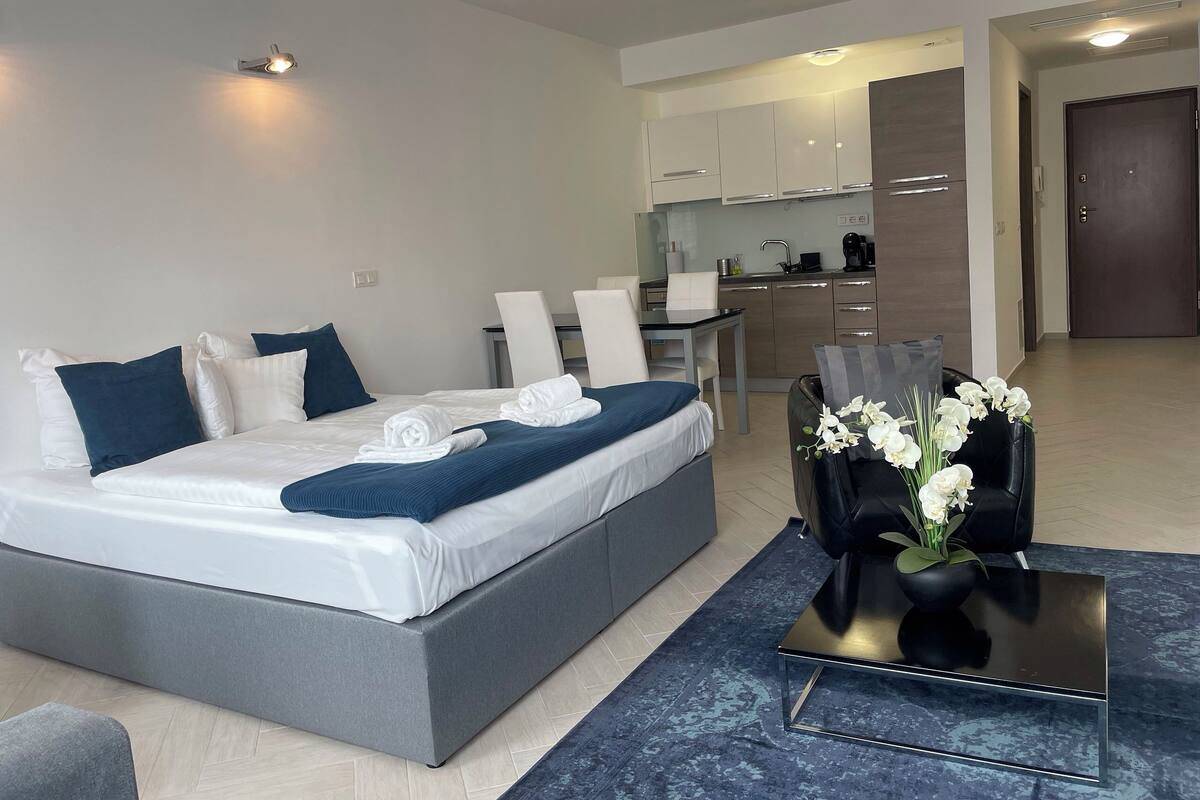 A modern hotel room with a large bed, a small dining table, a sofa, and a kitchen area.
