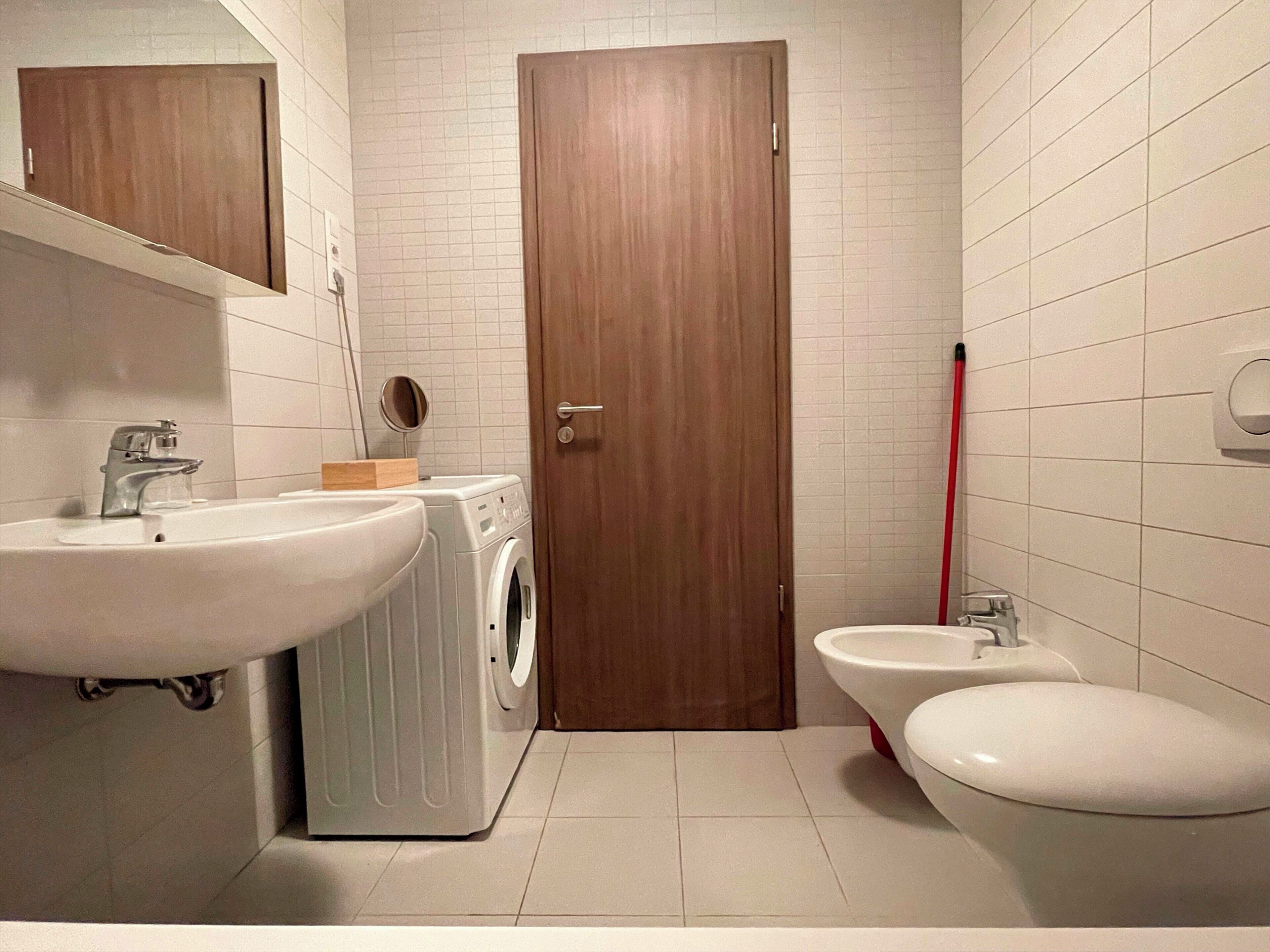 A bathroom with a sink, washing machine, toilet, and a wooden door.