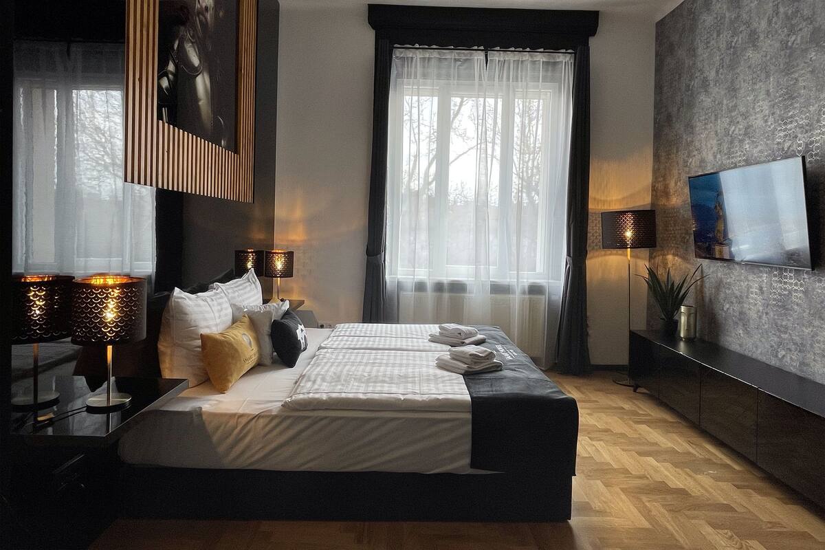 A modern bedroom with a large bed, bedside lamps, a TV, and a view of trees through the window.