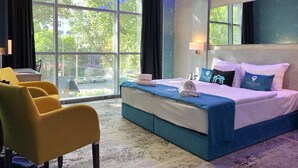 A hotel room with a large window, a bed with blue bedding, a yellow armchair, and a wall-mounted lamp. -  Smart Hotel Budapest (Budapest)