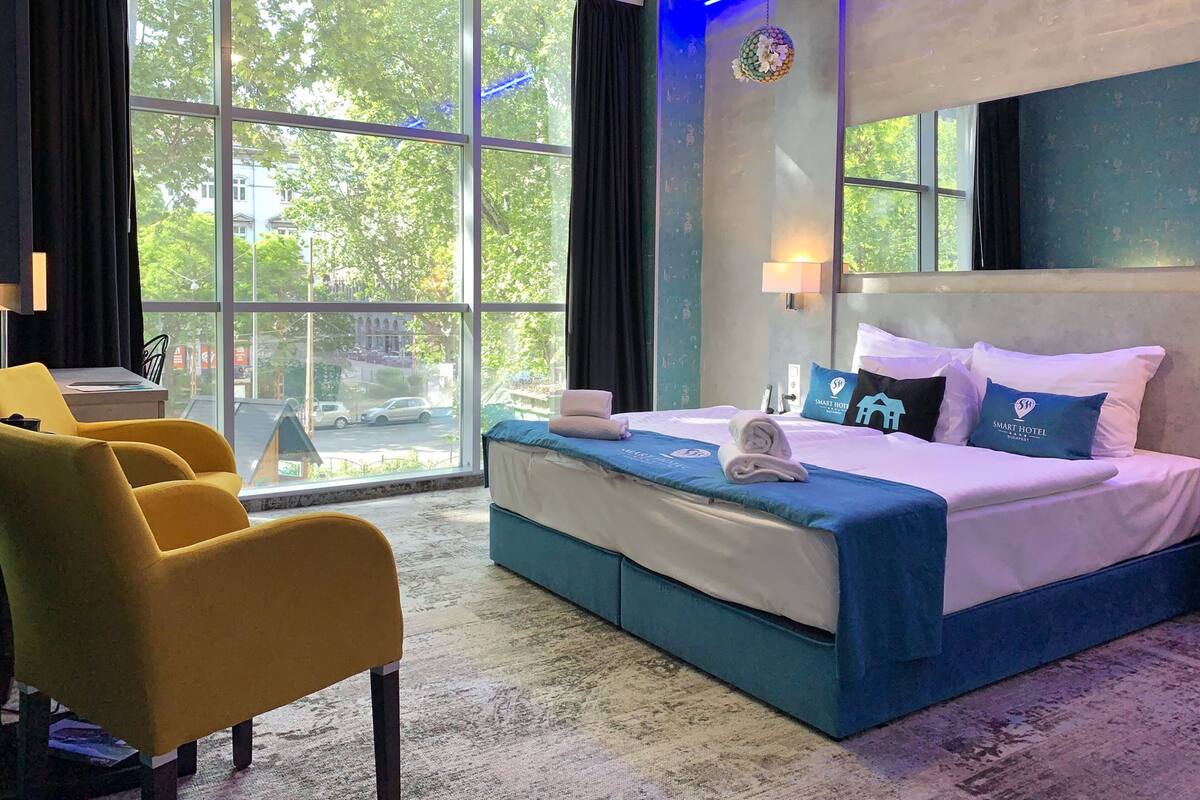 A hotel room with a large window, a bed with blue bedding, a yellow armchair, and a wall-mounted lamp.
