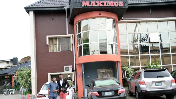 A building with a sign reading "MAXIMUS" and a curved entrance. There are three cars parked in front. - Maximus Suites (Lagos)