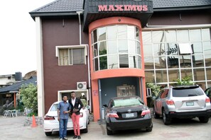 A building with a sign reading "MAXIMUS" and a curved entrance. There are three cars parked in front. - Maximus Suites (Lagos)
