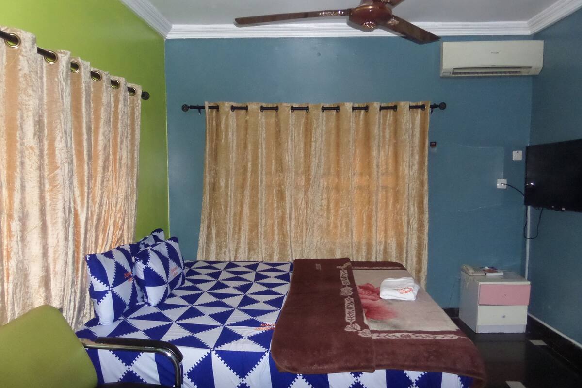 A room with a bed, a green chair, a small table with a blanket, a ceiling fan, an air conditioner, and a television.