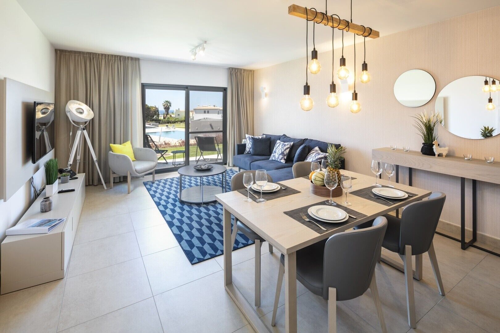 A modern hotel room with a dining area, a sofa set, a large window with a view, and a ceiling light fixture.