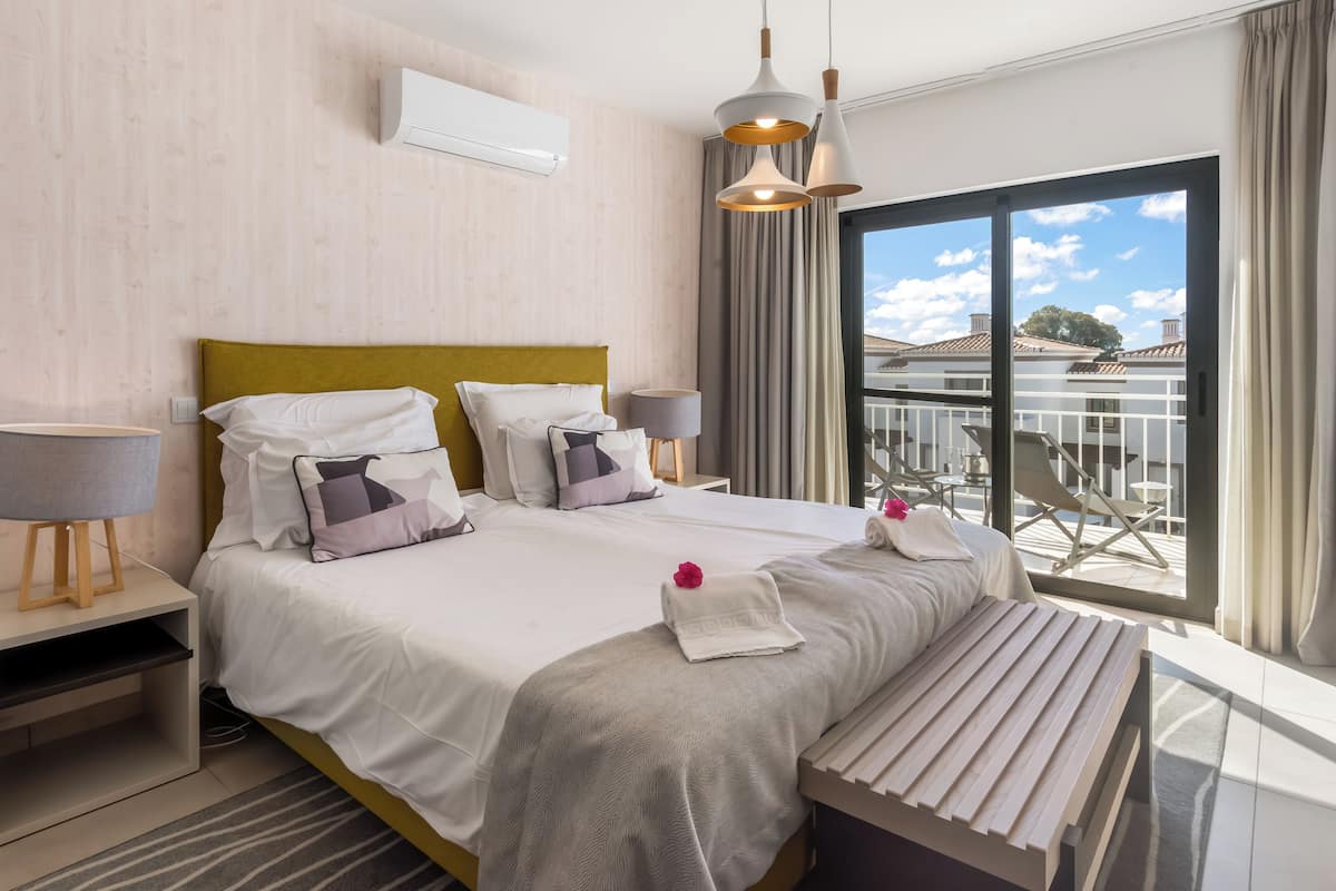 A modern hotel room with a large bed, bedside tables, a bench, and a balcony with a view.
