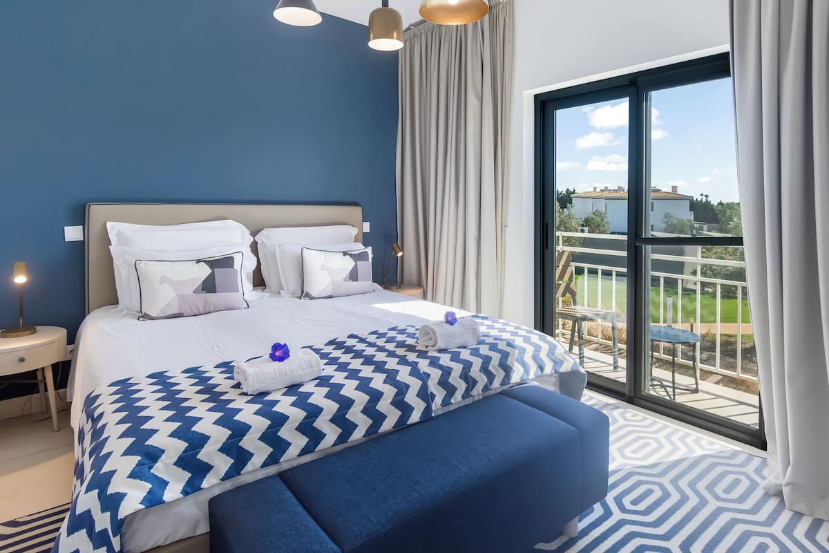 A hotel room with a large bed, a blue sofa, a balcony with a view, and a large window.