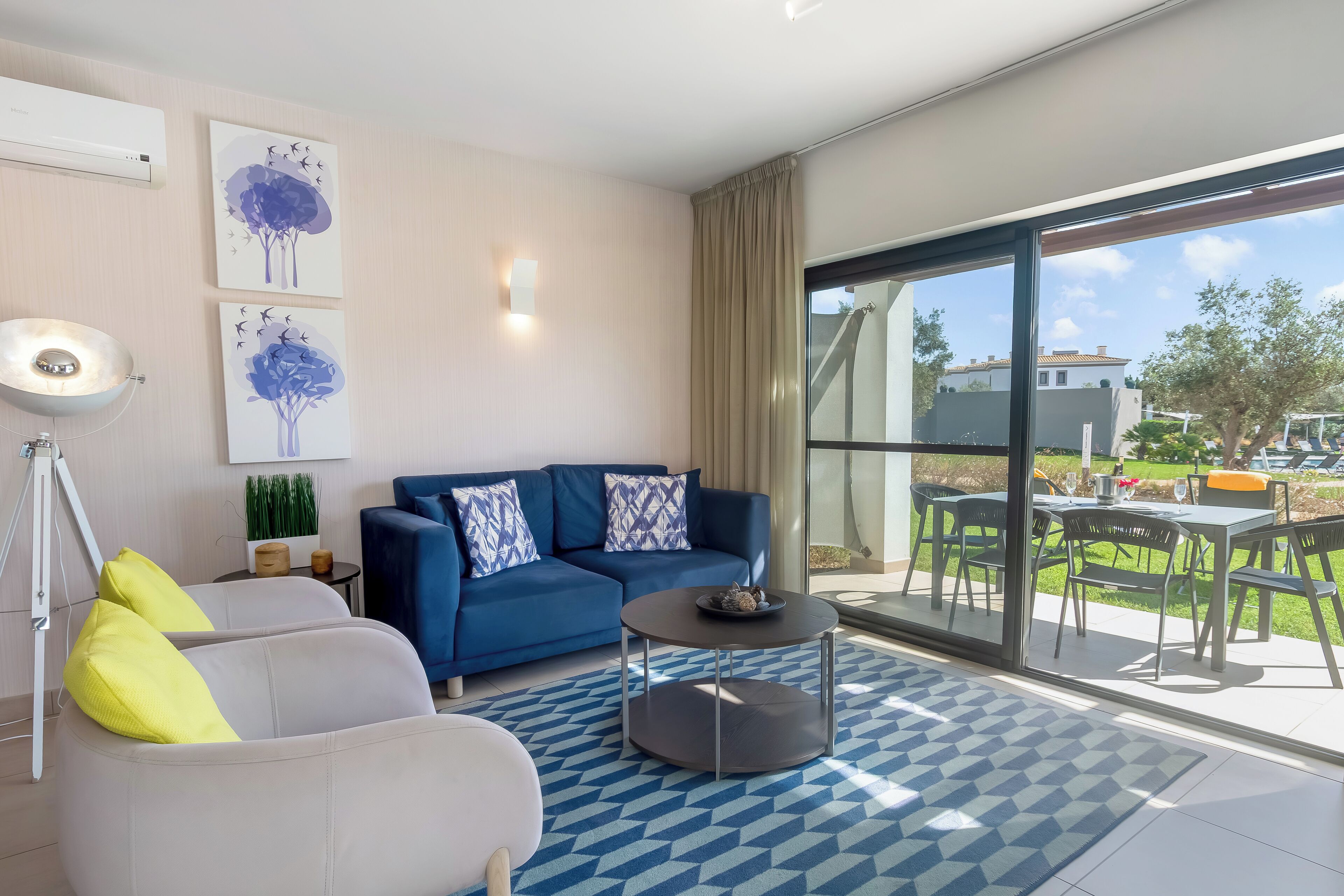 A modern living room with a blue sofa, white armchair, and a round coffee table. There is a large window with a view of the outdoors.