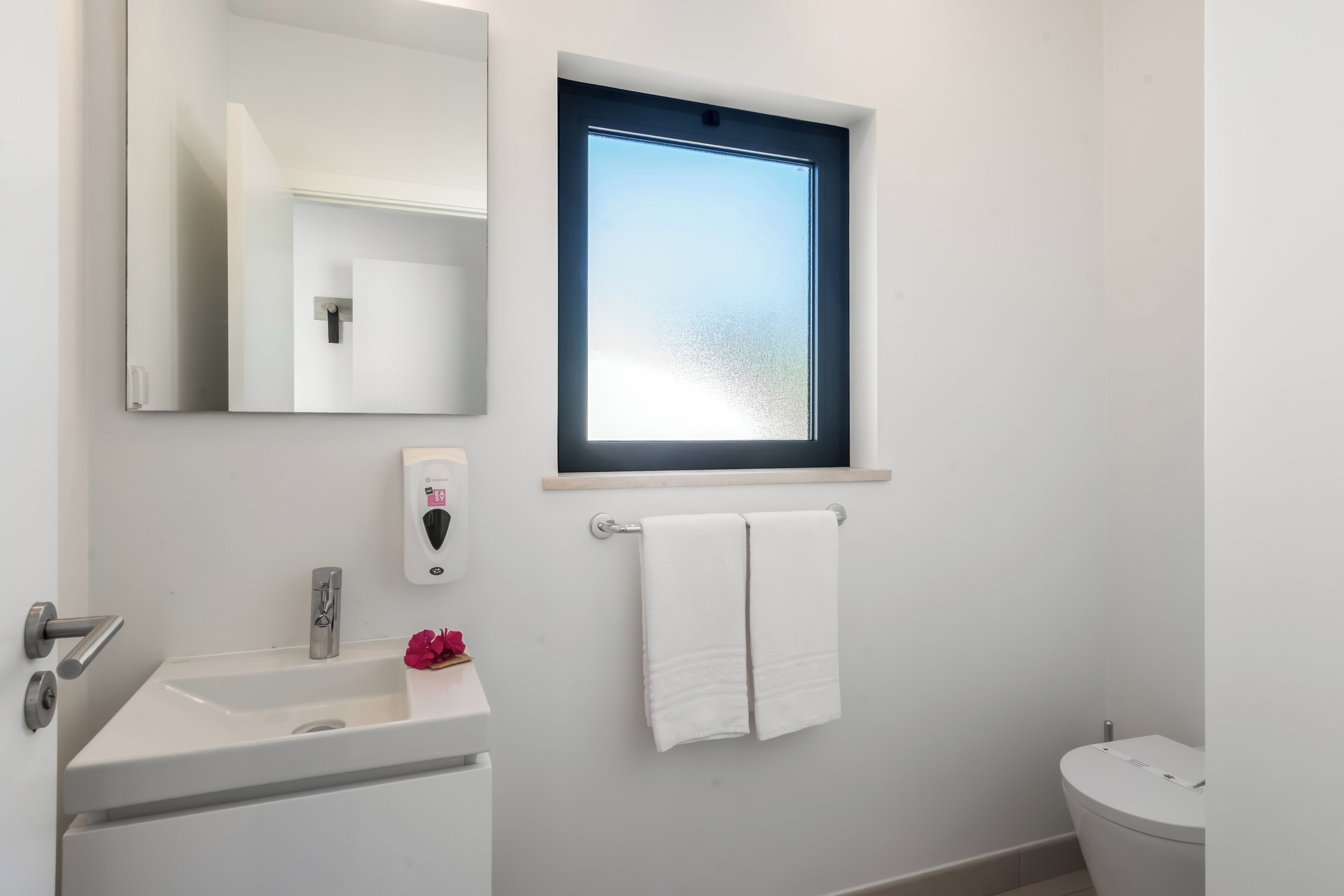 A bathroom with a white sink, a mirror, a window, and a toilet.