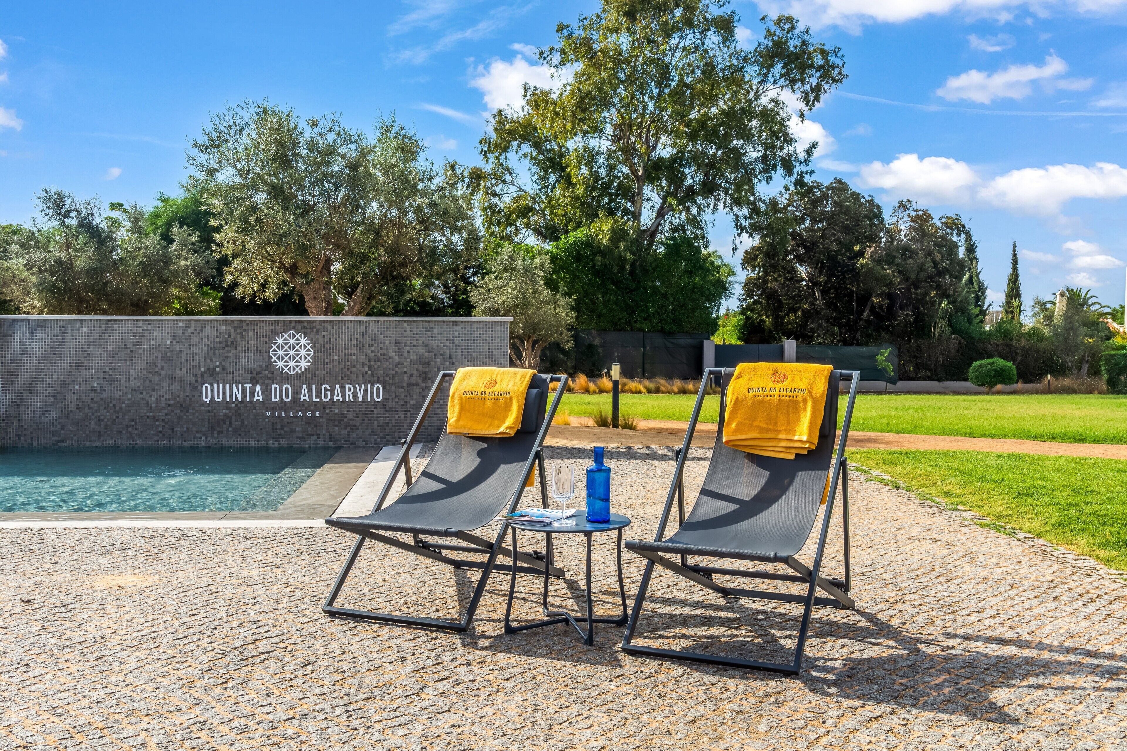 Deux chaises longues avec des serviettes jaunes, une petite table sur laquelle se trouve une bouteille bleue, et un espace piscine avec un mur carrelé portant l’inscription « QUINTA DO ALGARVIO VILLAGE ».