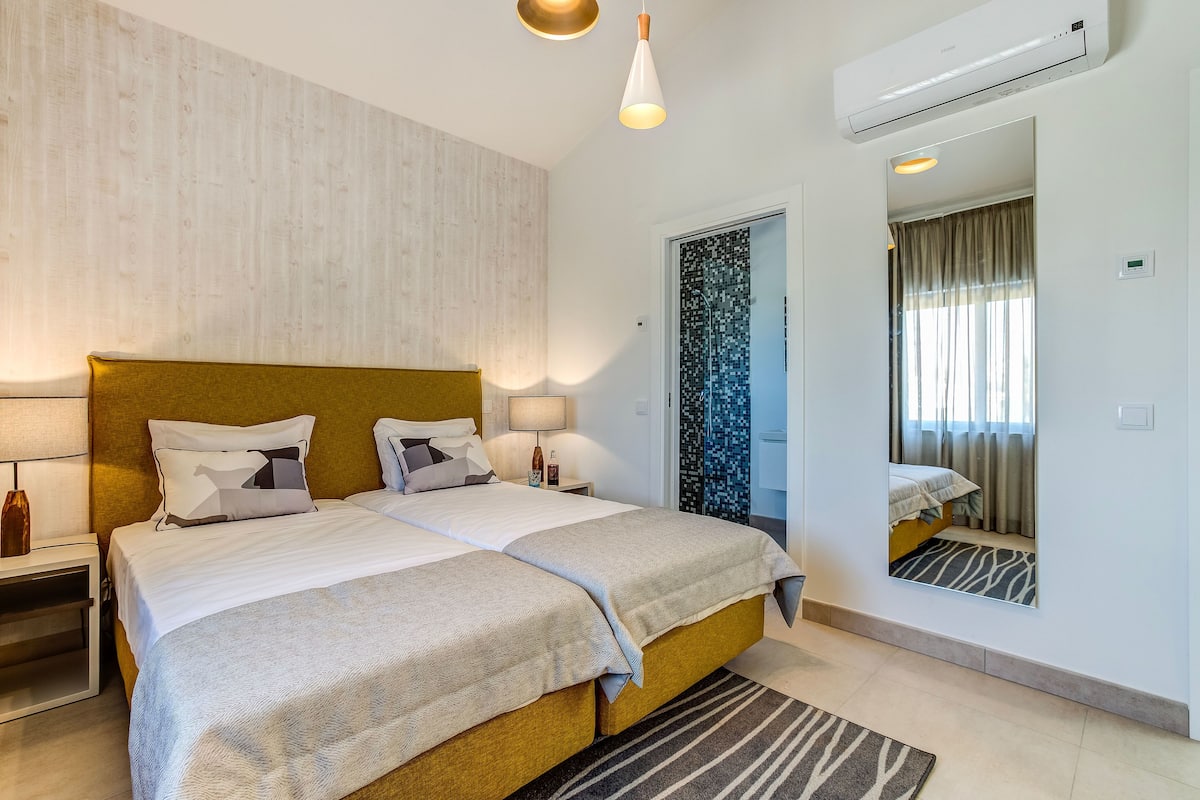 A double bed with a wooden headboard, bedside lamps, a mirror, and a view of a bathroom with a shower and curtains.