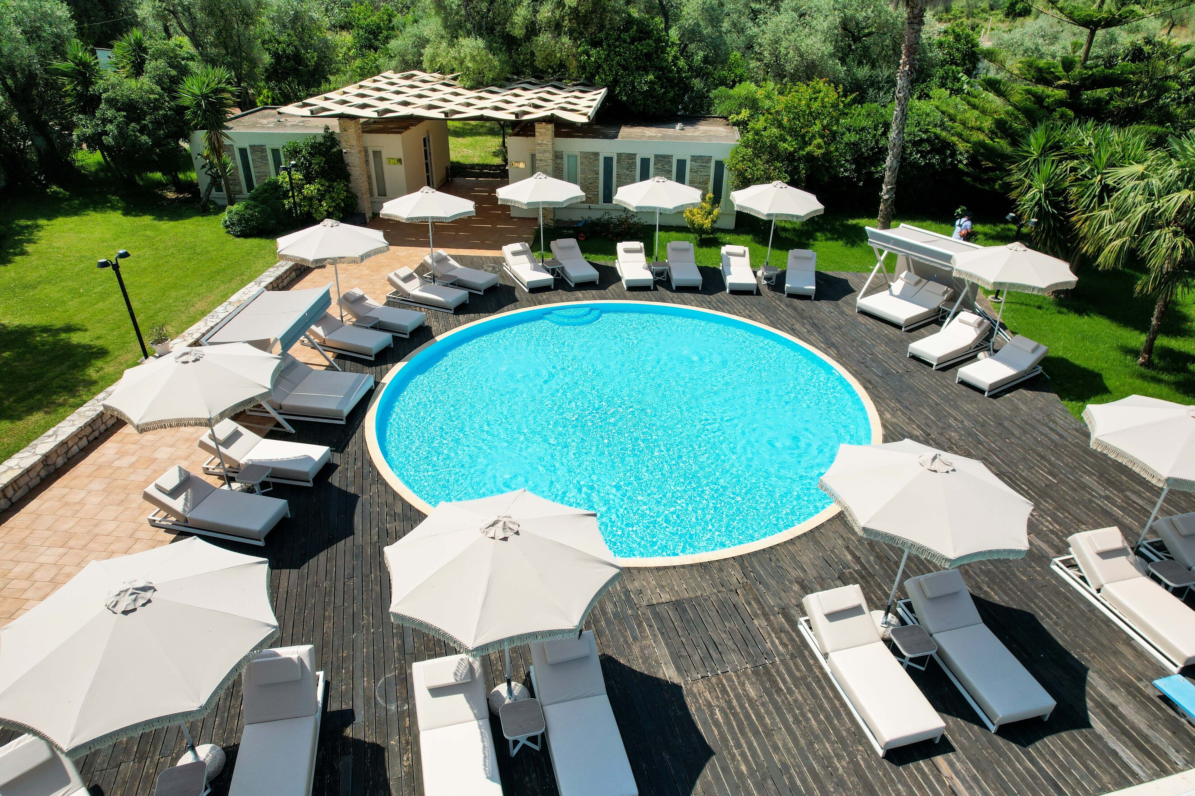A circular pool with lounge chairs and umbrellas, surrounded by a wooden deck and greenery.