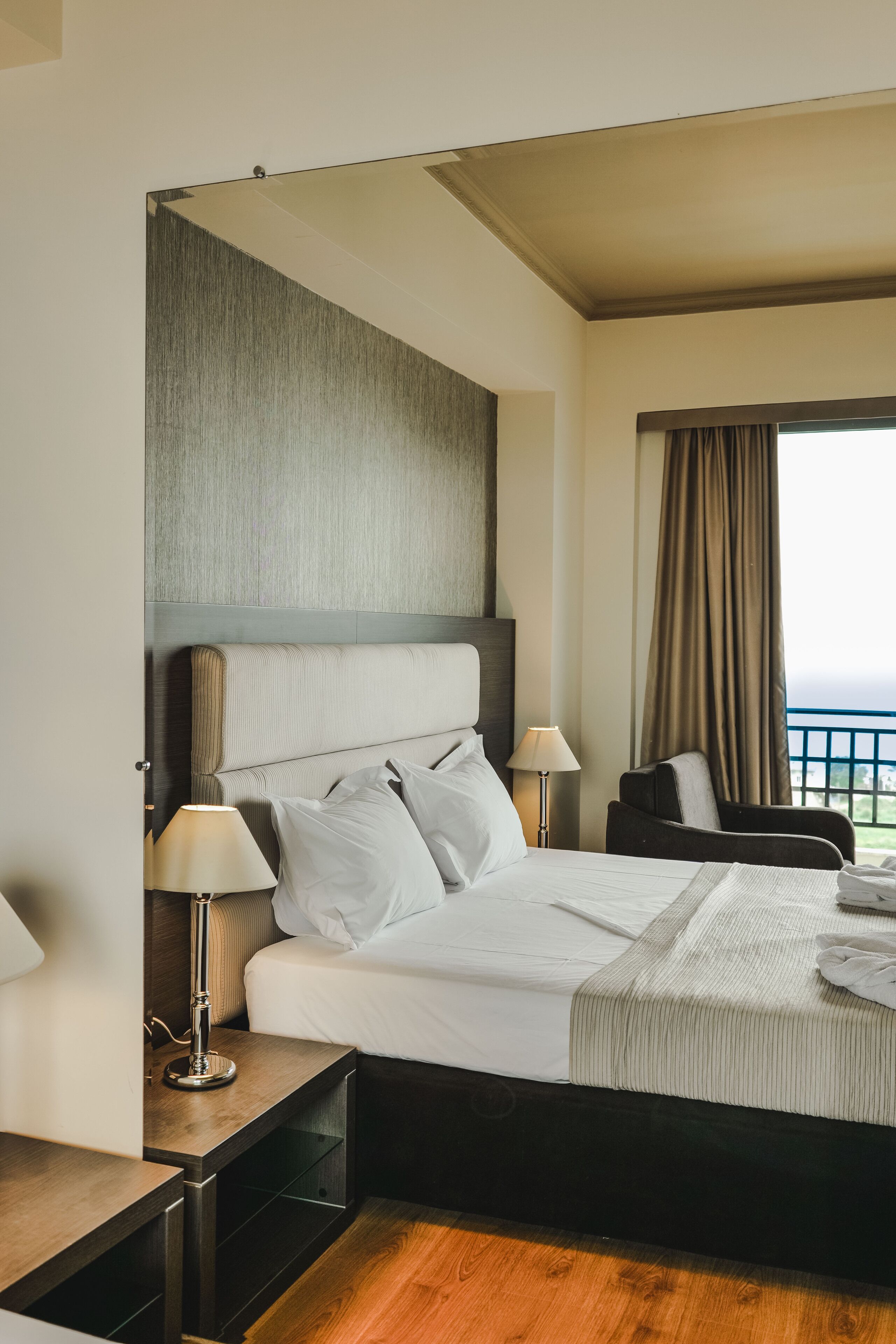 A hotel room with a large bed, two bedside tables with lamps, a chair, and a view of the sea through a window with curtains.