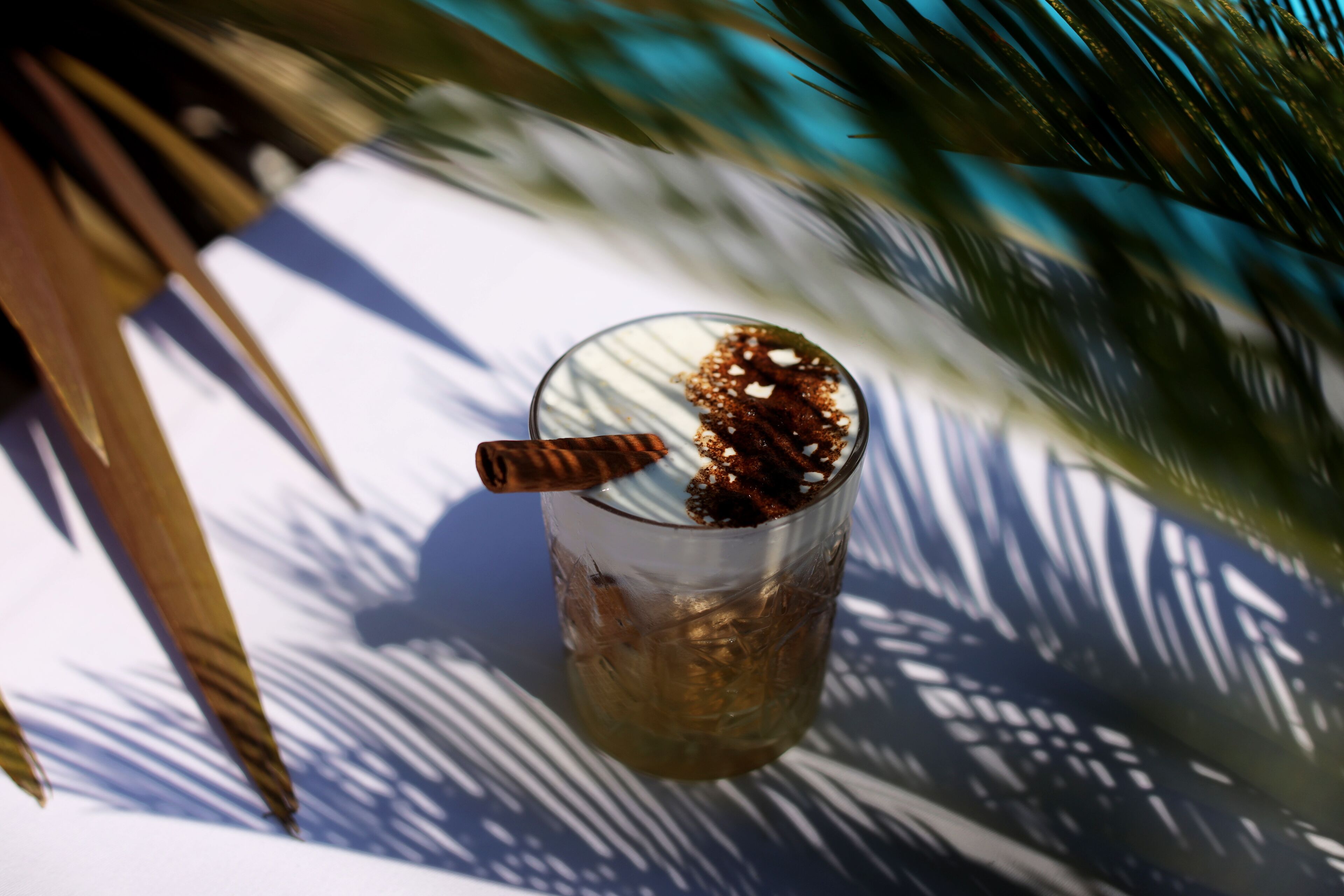 A glass with a straw and a spoon, placed on a surface with shadows of palm leaves.