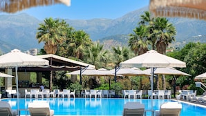 Un área de piscina con sillones de descanso y sombrillas, rodeada de palmeras y montañas al fondo.
